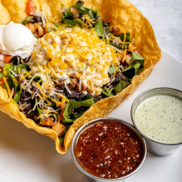 A taco salad in a crispy tortilla bowl with sour cream, salsa, and dressing on the side.