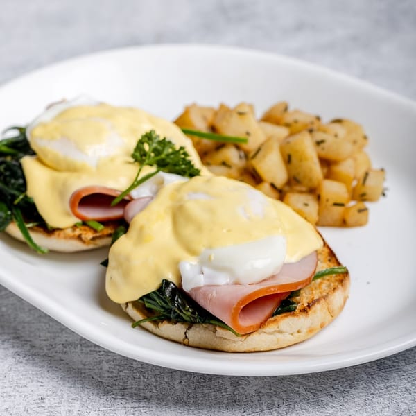 Two eggs Benedict with ham, spinach, hollandaise sauce, and a side of roasted potatoes.