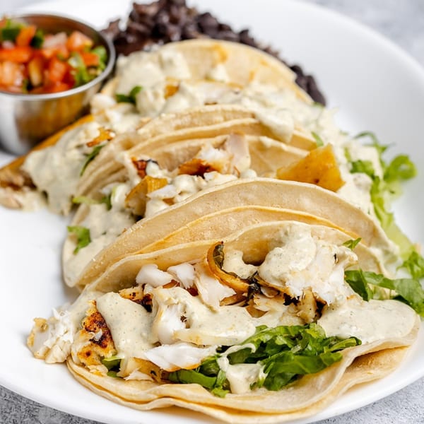 Four fish tacos with creamy sauce, lettuce, and black beans, served with a side of salsa.