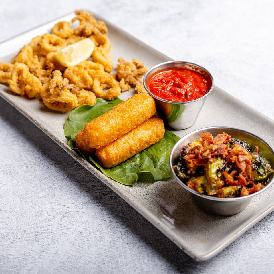 A platter of fried calamari, mozzarella sticks, roasted brussels sprouts with bacon, and marinara sauce.