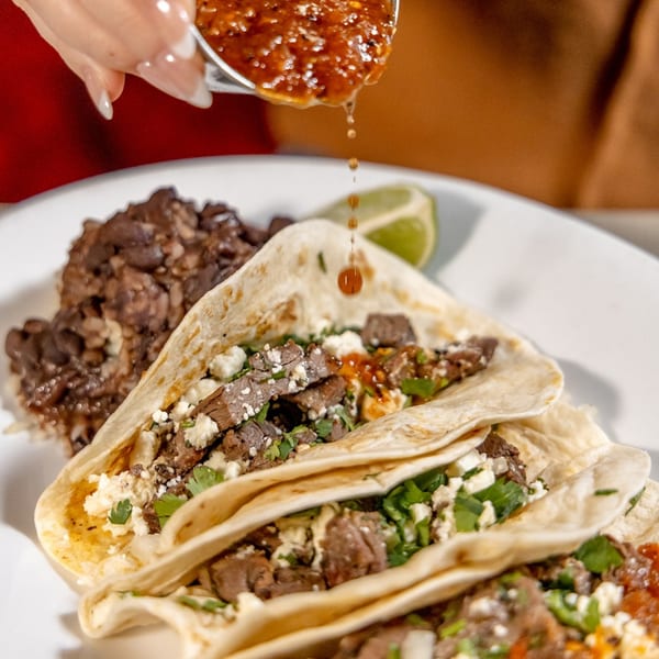 A hand drizzles red salsa over three steak tacos with crumbled cheese and cilantro, next to black beans.