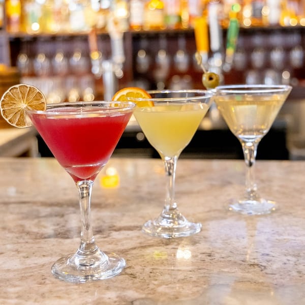 Three colorful cocktails, red, yellow, and clear, garnished with fruit and an olive, on a bar.
